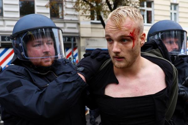 A young man with a bloody left eye is flanked by two German police officers in riot gear and helmets with face shields in the streets of Berlin.